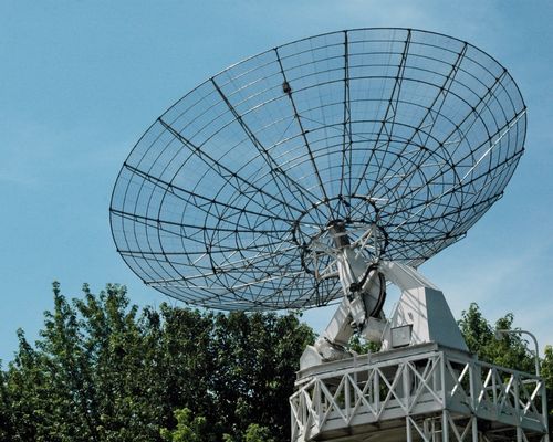 Large metal satellite dish with a mesh design, positioned above lush green trees under a clear blue sky