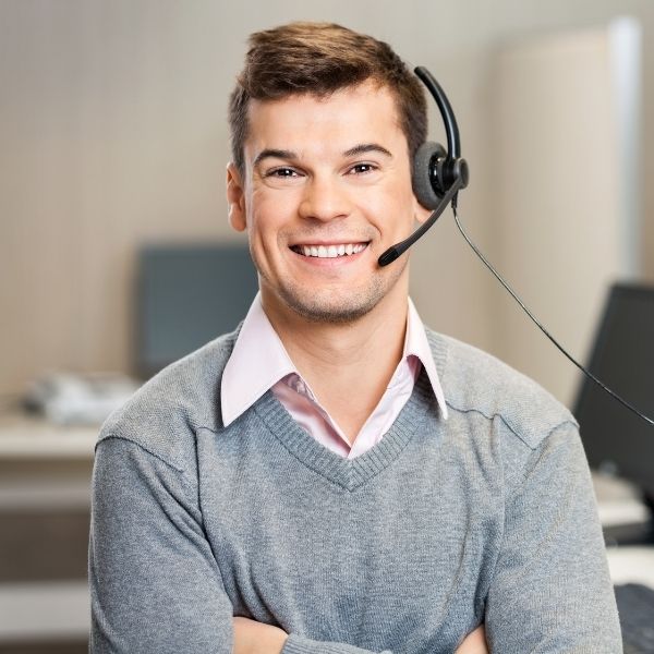 Smiling man wearing a gray sweater and pink shirt, with a headset, in an office setting